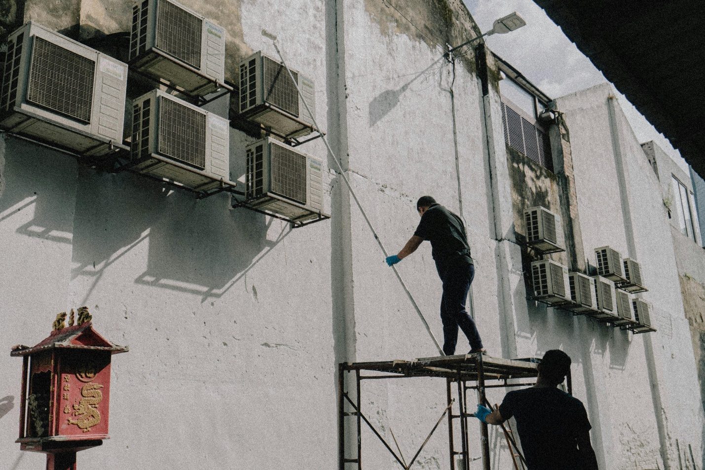 Two HVAC technicians on scaffolding servicing a wall of condenser units